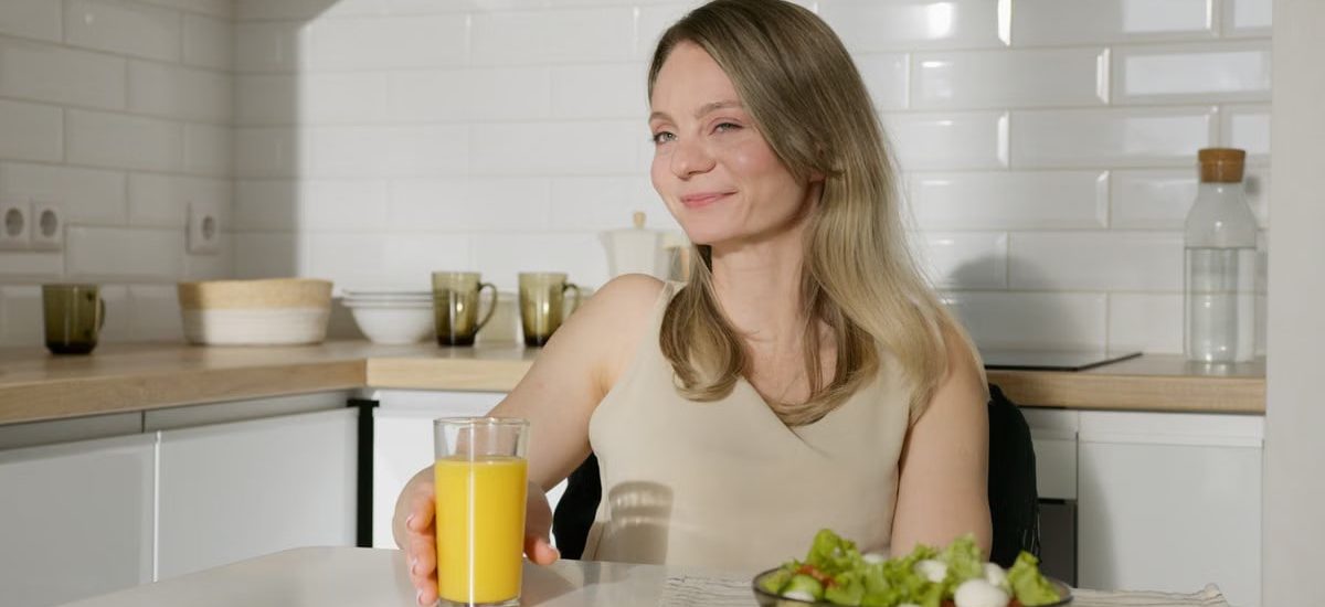 Smiling woman enjoying a healthy meal with salad and orange juice in a modern kitchen.