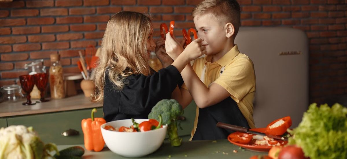 Playful boy and girl wearing apron preparing healthy vegetarian salad together at table with ingredients and bowls while looking at each other through slices of pepper