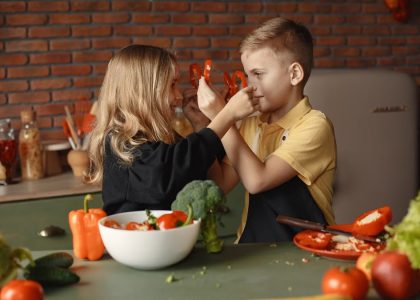 Playful boy and girl wearing apron preparing healthy vegetarian salad together at table with ingredients and bowls while looking at each other through slices of pepper